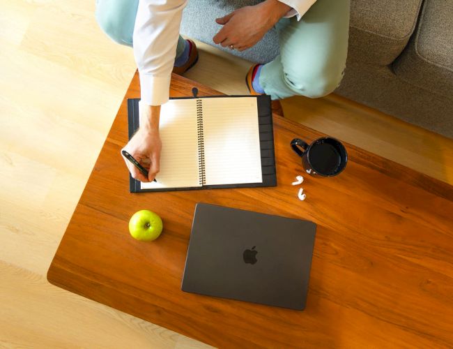 A person sits on a couch, writing in a notebook on a wooden table with a laptop, apple, coffee cup, scattered papers, and a few pills nearby.