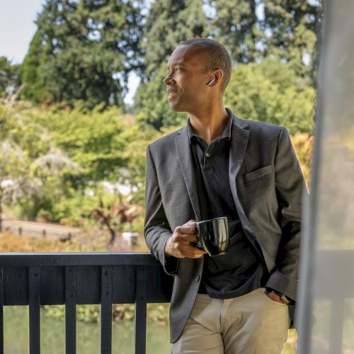 A person stands on a balcony holding a mug, gazing out at a lush, green landscape.