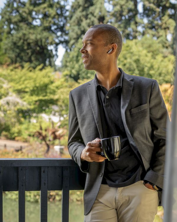 A person stands on a balcony holding a mug, gazing out at a lush, green landscape.