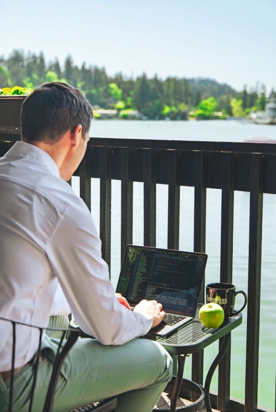 A man in a white shirt sits at a small table on a balcony by the water, typing on a laptop with an apple and mug nearby.