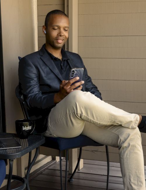 A person is sitting on a chair, looking at a phone. There's a table with a laptop and mug nearby on a balcony or patio.