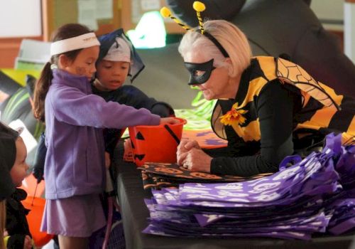 Children in costumes are trick-or-treating, receiving candy from an adult dressed as a bee in a Halloween setting.
