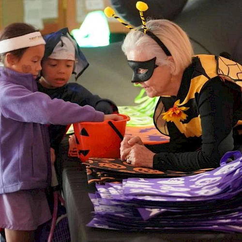 Children in costumes are trick-or-treating, receiving candy from an adult dressed as a bee in a Halloween setting.