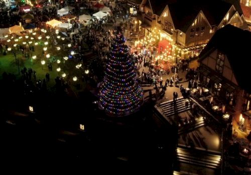 Aerial view of a festive, illuminated Christmas market with a large decorated tree, booths, and a crowd of people enjoying the event.
