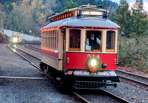 A vintage red streetcar adorned with festive lights travels on railway tracks through a wooded area. A sign reads "Lake Oswego."