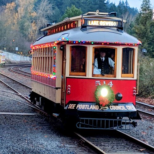 A vintage red streetcar adorned with festive lights travels on railway tracks through a wooded area. A sign reads "Lake Oswego."