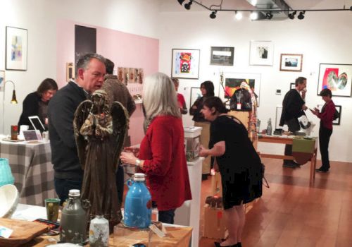 People are in an art gallery, observing and discussing various artworks displayed on the walls and tables.