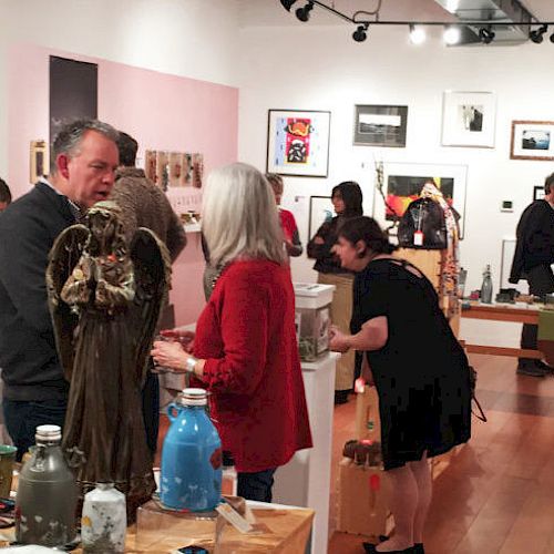 People are in an art gallery, observing and discussing various artworks displayed on the walls and tables.