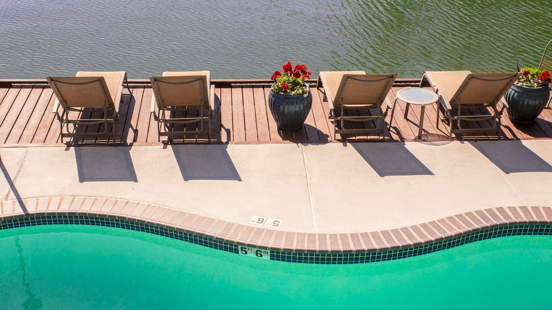Lounge chairs and small tables beside a curved pool, overlooking a body of water with planters placed along the deck.