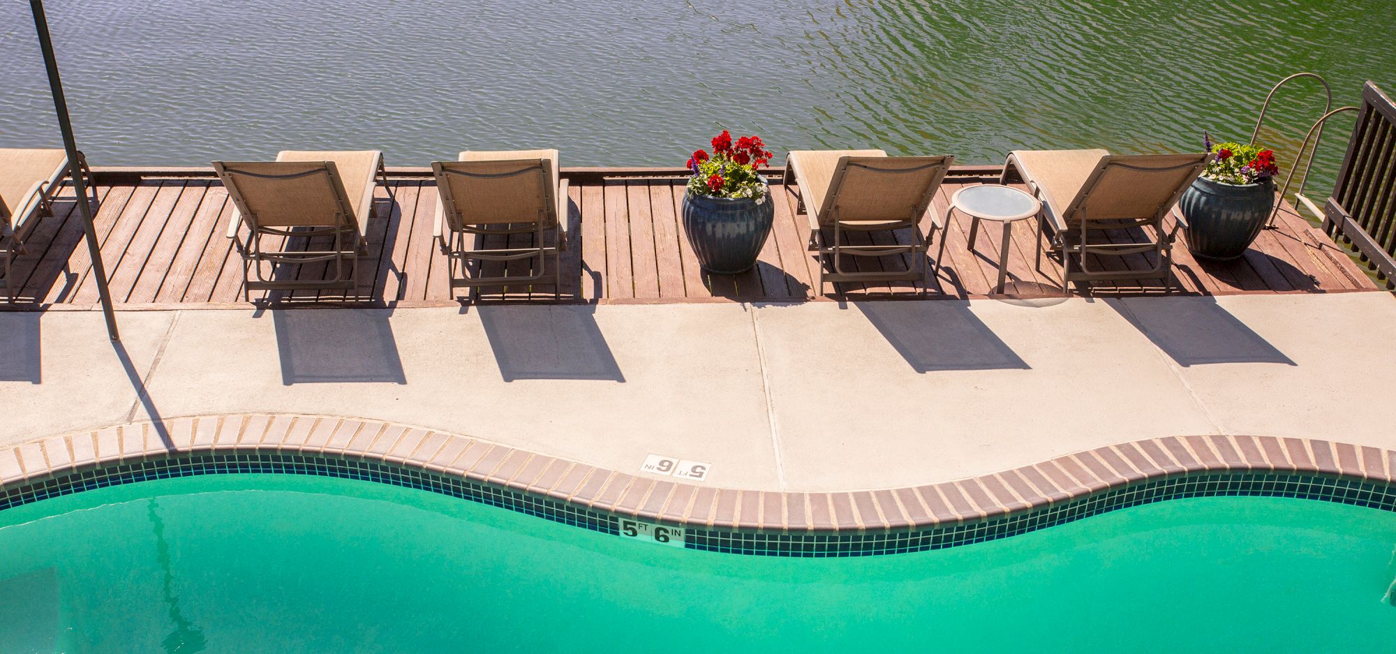 Lounge chairs and small tables beside a curved pool, overlooking a body of water with planters placed along the deck.