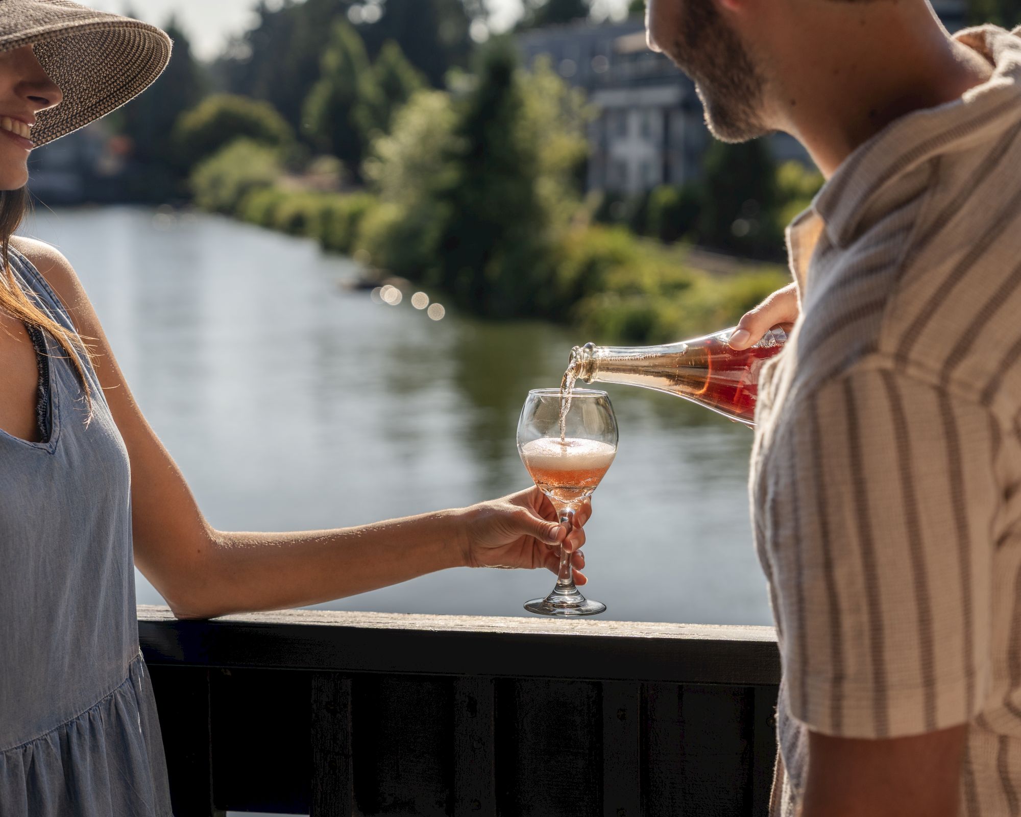 Two people clink glasses by a railing over a lake, outdoors on a sunny day.