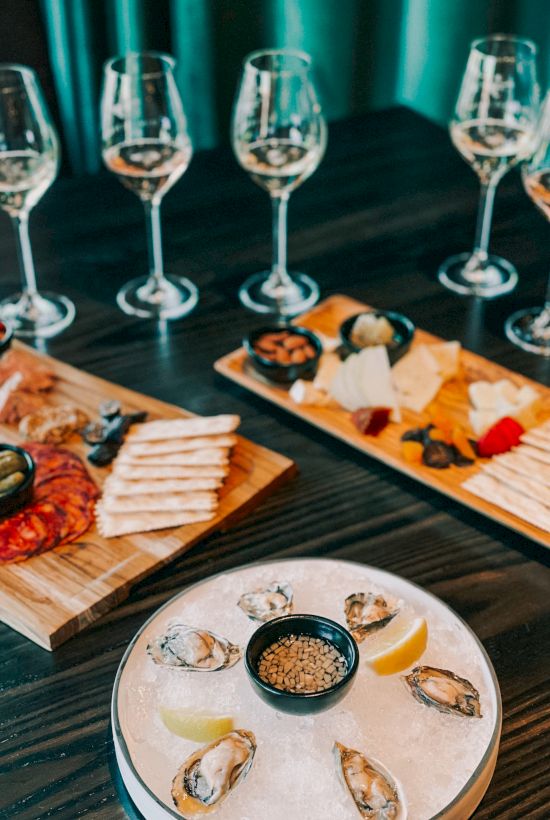 A tasting spread with wine glasses and boards of sushi, charcuterie, and oysters, set on a dark table against teal curtains, elegant and inviting.