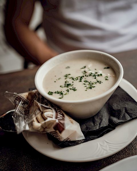 Creamy soup in a white bowl topped with herbs, served with a wrapped pastry on a black napkin, on a white plate.