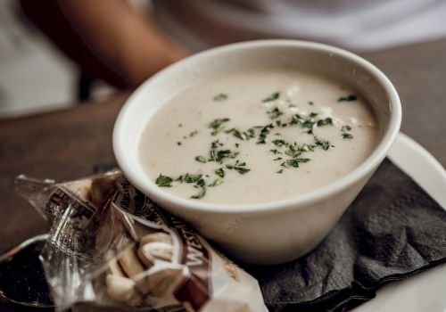 Creamy soup in a white bowl topped with herbs, served with a wrapped pastry on a black napkin, on a white plate.