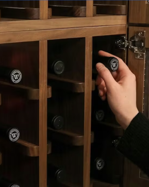 A person is placing or removing a bottle from a wooden wine rack with individual slots. It&rsquo;s a close-up of hands handling a bottle.