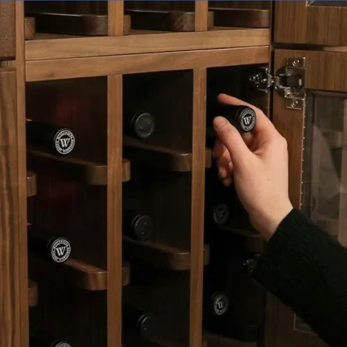 A person is placing or removing a bottle from a wooden wine rack with individual slots. It&rsquo;s a close-up of hands handling a bottle.