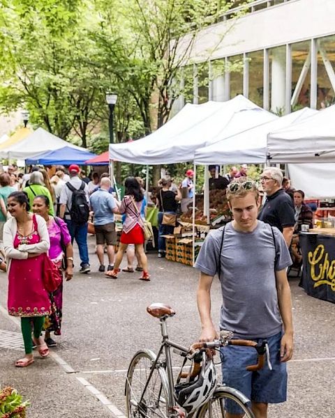 A lively outdoor market scene with stalls, people strolling, a man with a bicycle, and colorful vendor tents along a tree-lined street.