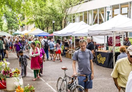 A lively outdoor market scene with stalls, people strolling, a man with a bicycle, and colorful vendor tents along a tree-lined street.