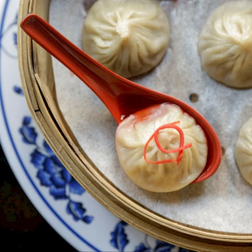 Steamed dumplings in a basket with one lifted by a red spoon, showing a small pink garnish on top, on a blue-patterned plate.