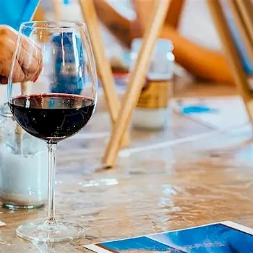 People painting at a table, sipping wine, with brushes, canvases, and photos scattered around.