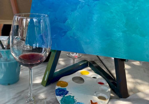 A painting setup outdoors with a blue canvas on an easel, a wine glass, and a painter&rsquo;s palette with mixed paints nearby.