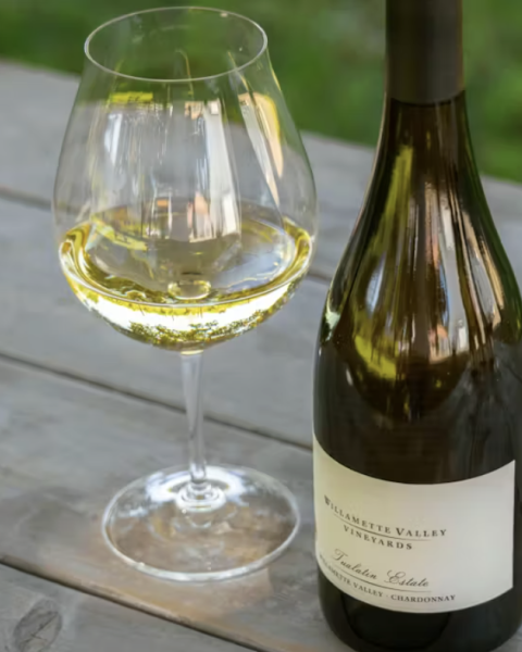 A glass of white wine beside a bottle on a wooden table, outdoors on a sunny day.