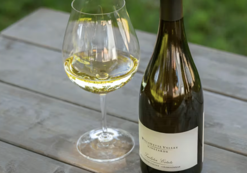 A glass of white wine beside a bottle on a wooden table, outdoors on a sunny day.