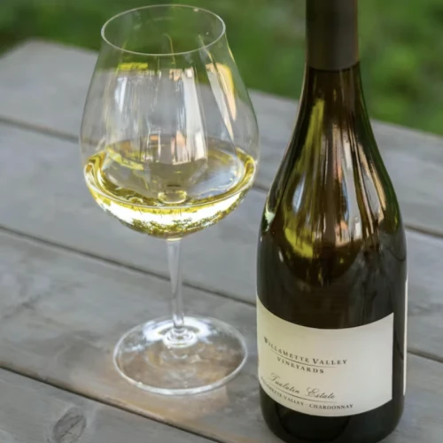 A glass of white wine beside a bottle on a wooden table, outdoors on a sunny day.