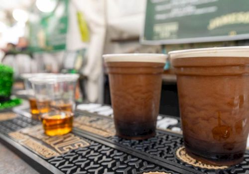 A row of coffee cups on a bar counter with a chalkboard menu in the blurred background, plus a glass of whiskey on the side.