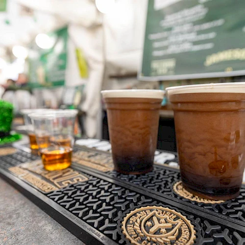 A row of coffee cups on a bar counter with a chalkboard menu in the blurred background, plus a glass of whiskey on the side.