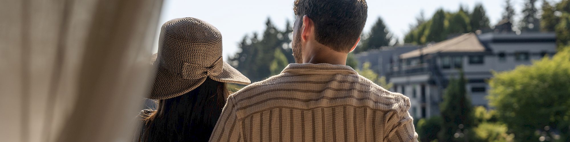 A couple stands on a balcony overlooking a serene view, with one holding a glass of wine, framed by a curtain.