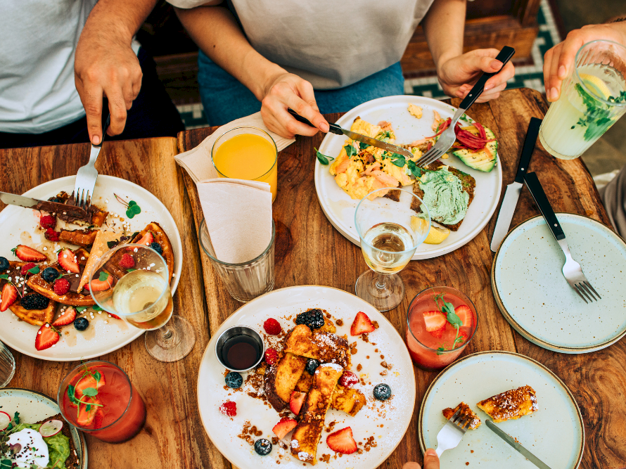 A table full of colorful plates with brunch items and drinks, people sharing food, hands reaching for utensils and plates, sunny dining vibe.