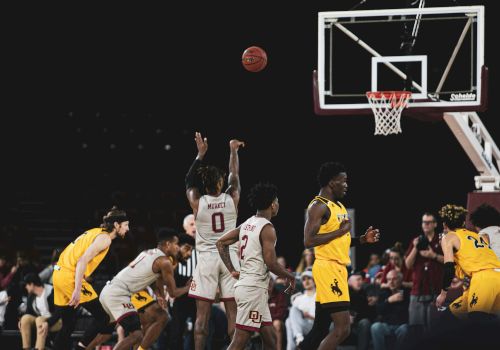 A basketball game in progress; players jump for a shot near the basket as others guard and spectators watch intently.