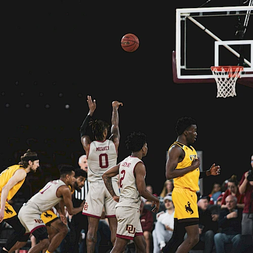 A basketball game in progress; players jump for a shot near the basket as others guard and spectators watch intently.