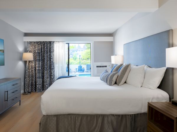 Modern hotel bedroom with a bed, blue patterned curtains, a sliding door to the outdoors, bedside lamps, and an air conditioner.
