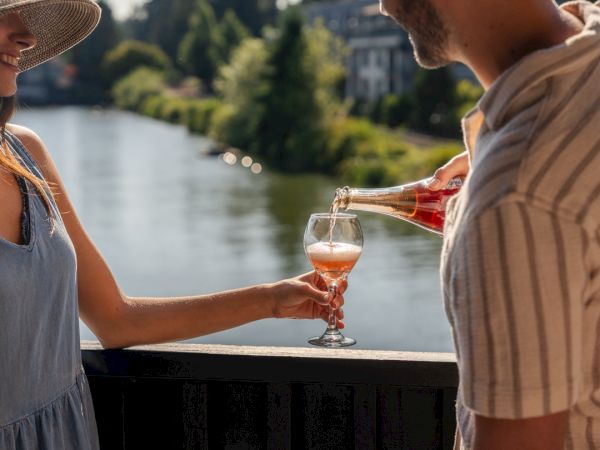 Two people clink wine glasses on a sunlit waterfront deck, smiling as they toast near a serene lake.