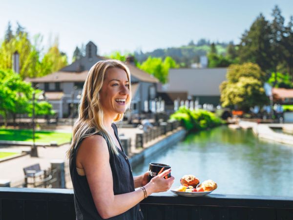 A smiling woman stands on a balcony by a sunny canal, holding a coffee cup and a plate of pastries with a scenic waterfront backdrop.