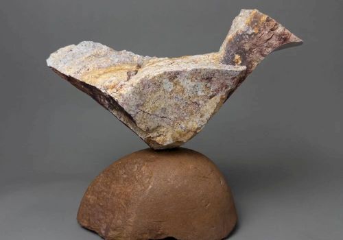 A sculpture of a stylized bird perched on a rounded pedestal, made from rough, layered clay or stone with a weathered finish.
