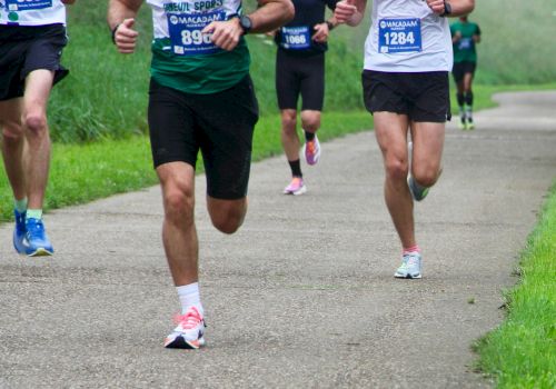 A group of runners in a race on a paved path, wearing numbered bibs and athletic gear, mid-stride on a grassy trail.