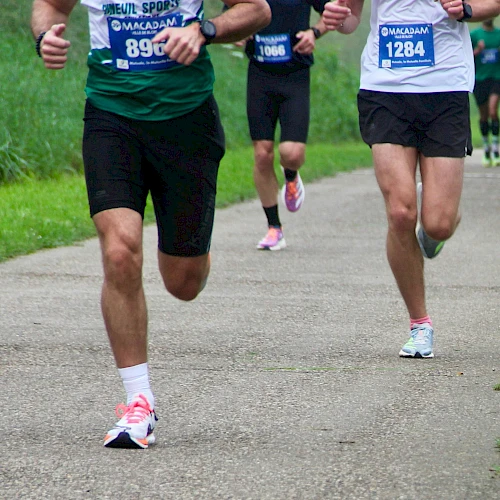 A group of runners in a race on a paved path, wearing numbered bibs and athletic gear, mid-stride on a grassy trail.