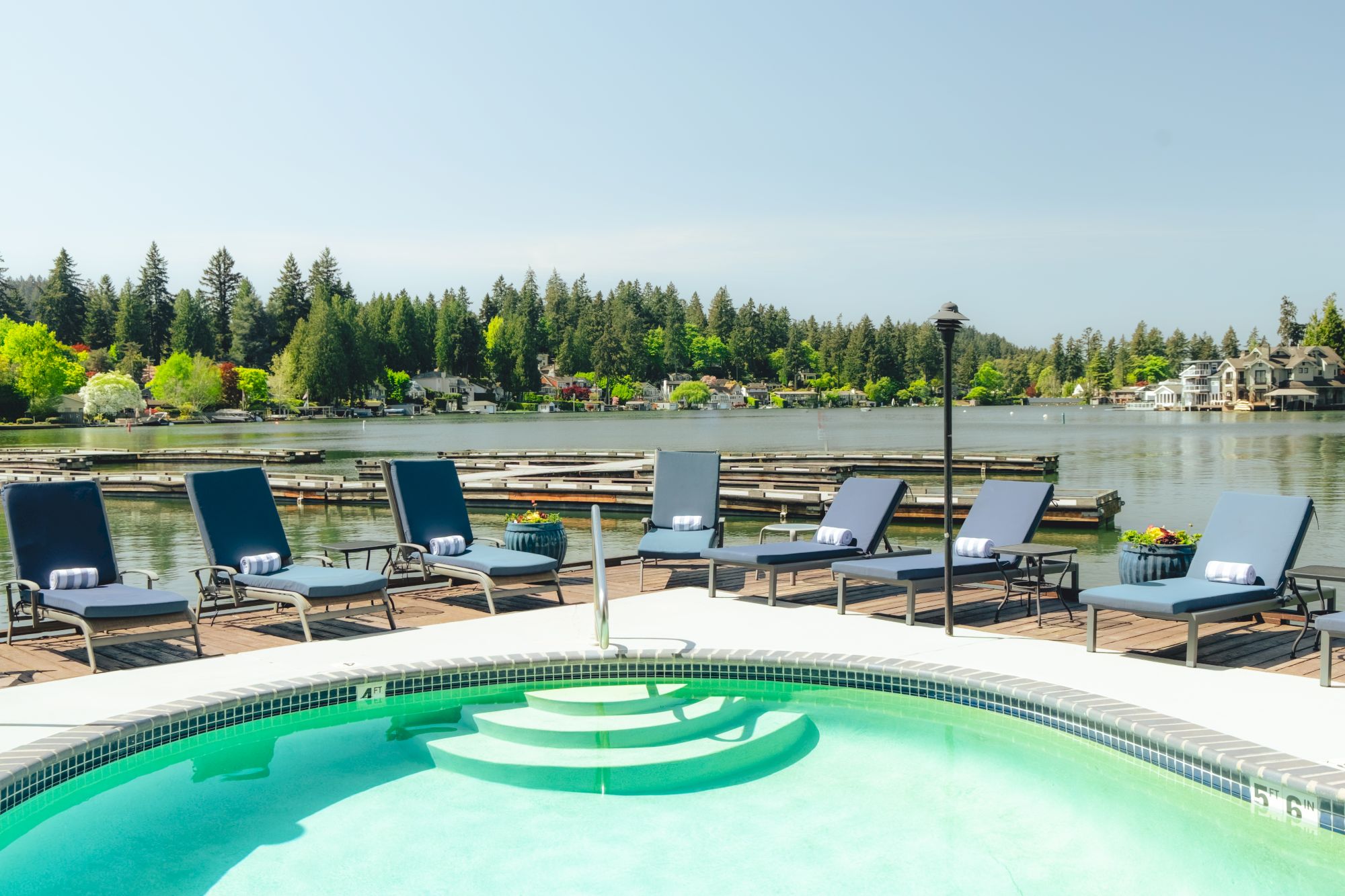 A lagoon-style pool with bubbling turquoise water, a circular hot tub in the foreground, and a row of blue lounge chairs along a dock by a serene lake, surrounded by trees.