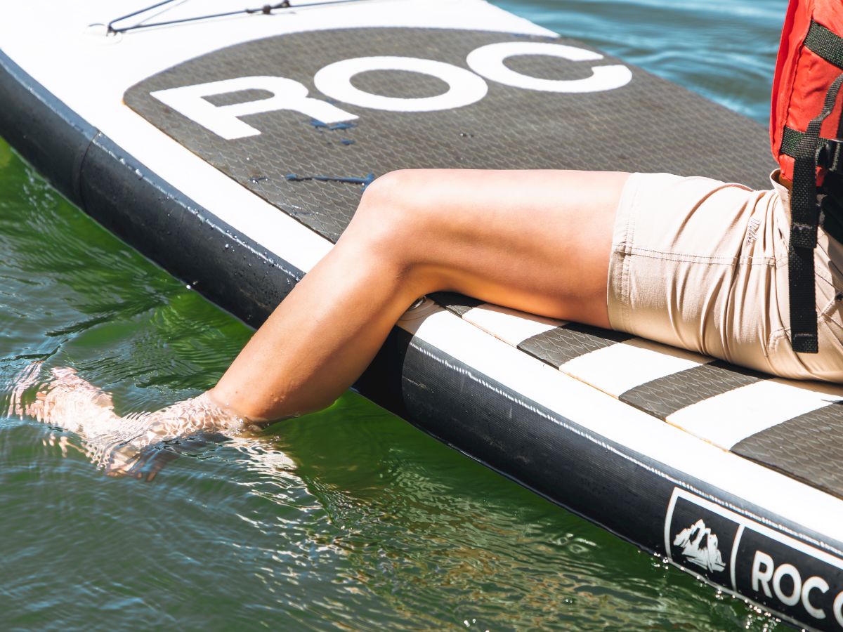 A person sits on a paddleboard in a lake, wearing a life vest, with legs resting on the board and water lapping at the edge.