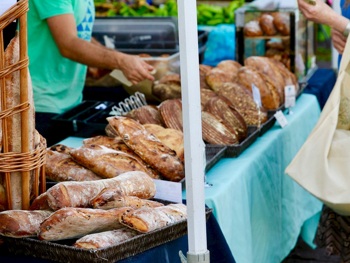 Fresh bread and loaves on a market stall, with customers browsing and a basket visible, local bakery vibes.