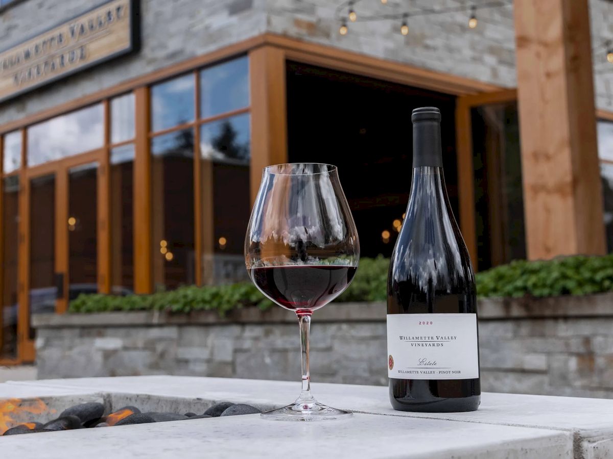 A glass of red wine and a bottle sit on a table outside a rustic cafe or restaurant, with warm lights and stone/wood decor in the background.