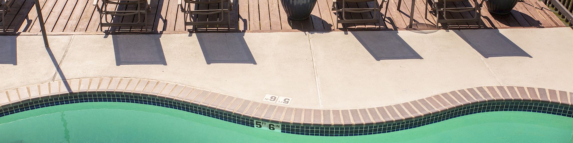 A sunny poolside scene with lounge chairs, planters, and a curved pool by the water, on a bright day.