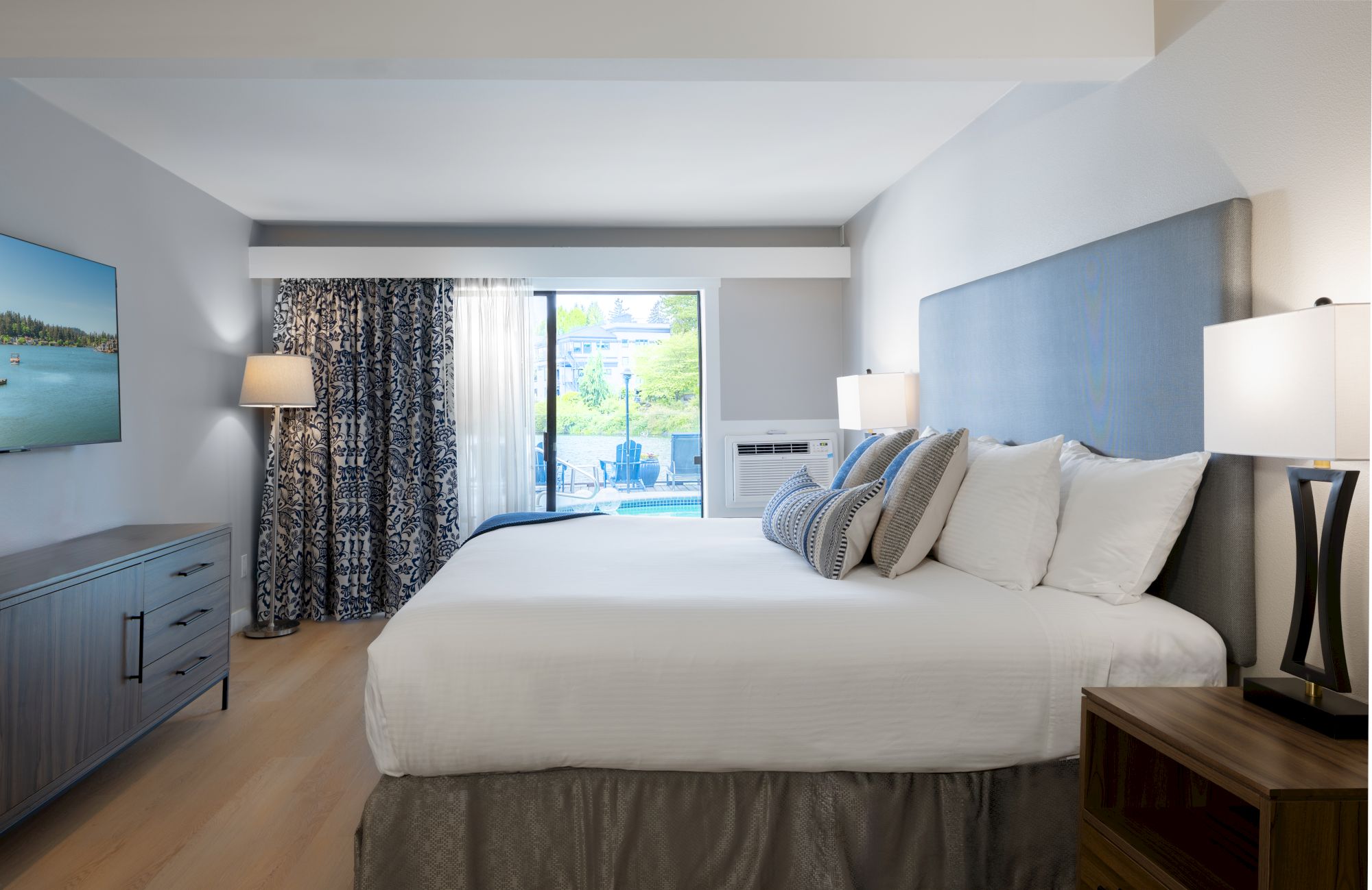 A modern hotel room with a large bed, white linens, blue headboard, two nightstands with lamps, a dresser, and a sliding door to a balcony.