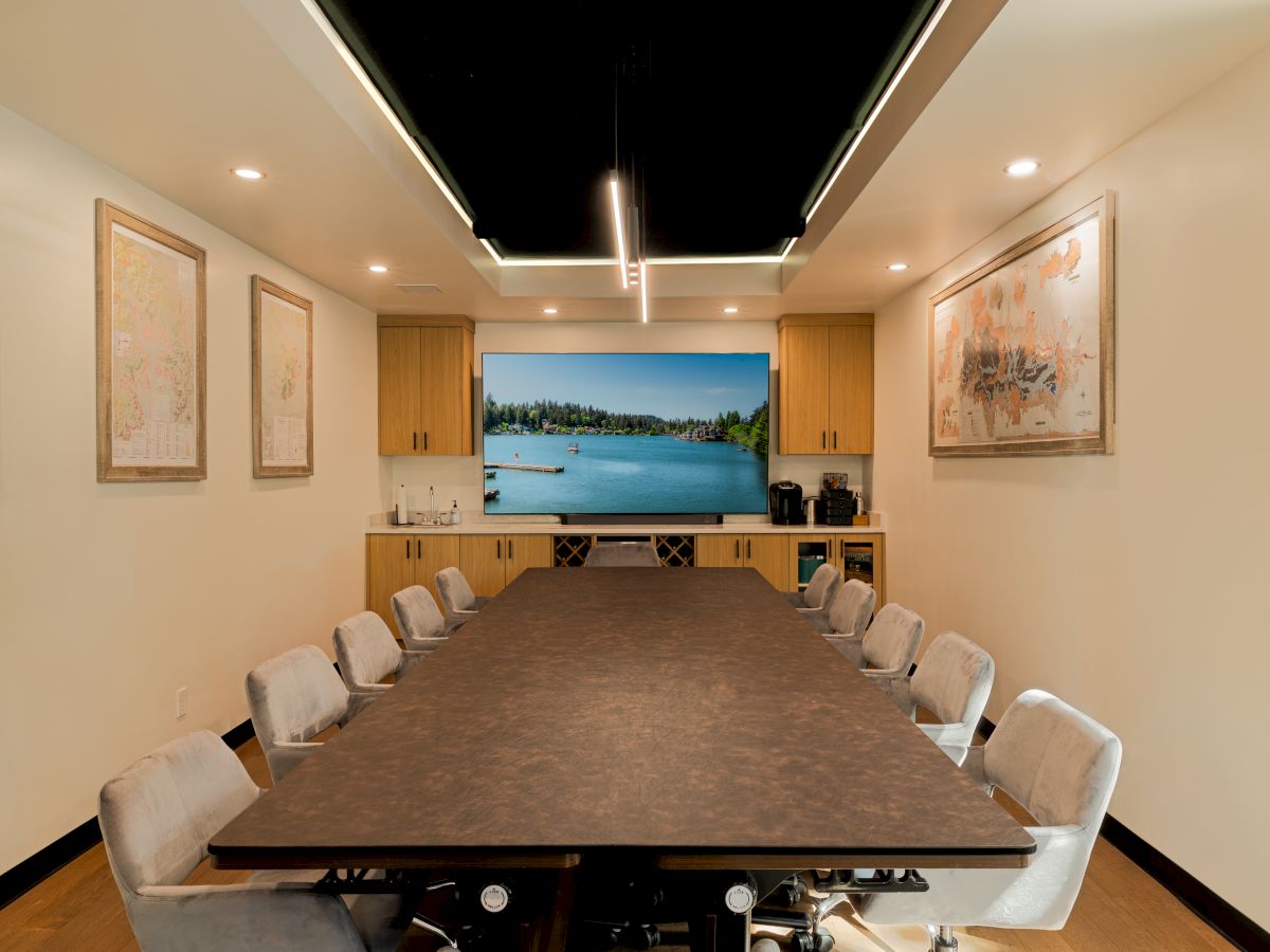 A long conference room with a central table, white chairs, wall maps/art, and a large screen at the far end showing a beach scene to inspire.