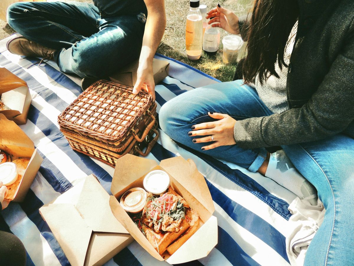 A cozy picnic scene: two people on a striped blanket with food boxes, a woven basket, drinks, and snacks all laid out together.