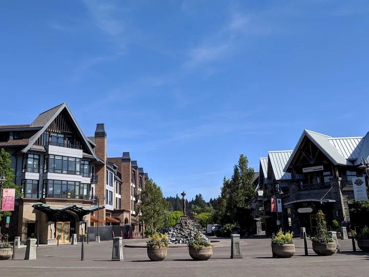 A quaint alpine village square with timber-framed buildings, outdoor shops, planters, and a clear blue sky.
