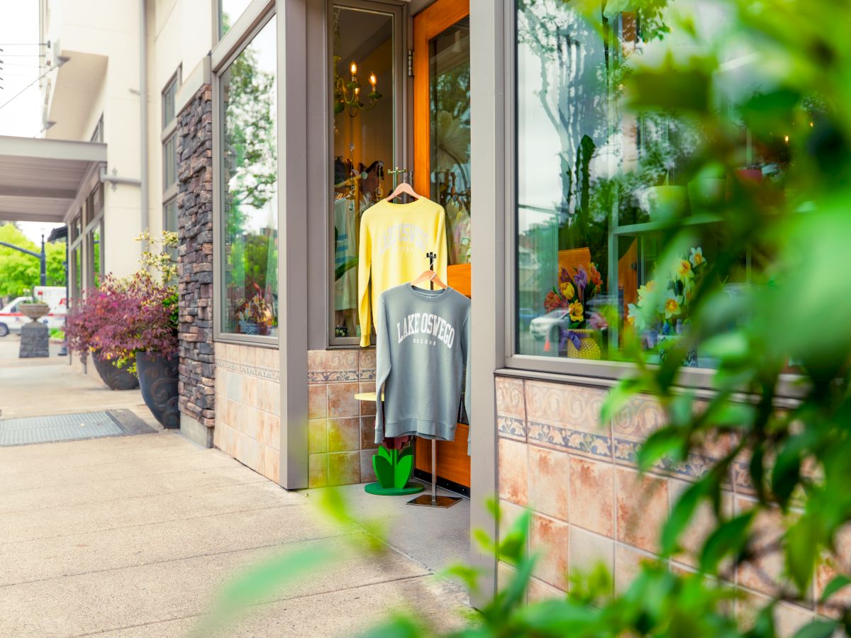 A storefront window with two t‑shirts on display outside, one yellow and one gray, on a sunny sidewalk with green plants nearby.